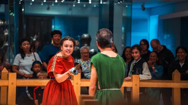 Two reenactors in red and green tunics perform a mock sword fight with wooden weapons inside a fenced area, while spectators observe from behind
