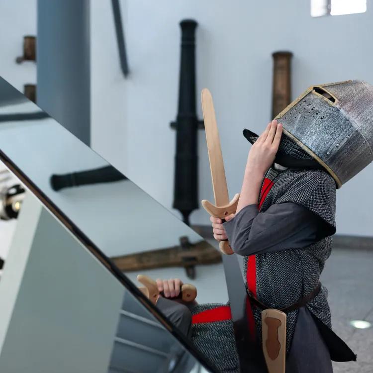 child dressed as a knight gazes upwards in front of a mirror reflecting the interior of the hall of steel