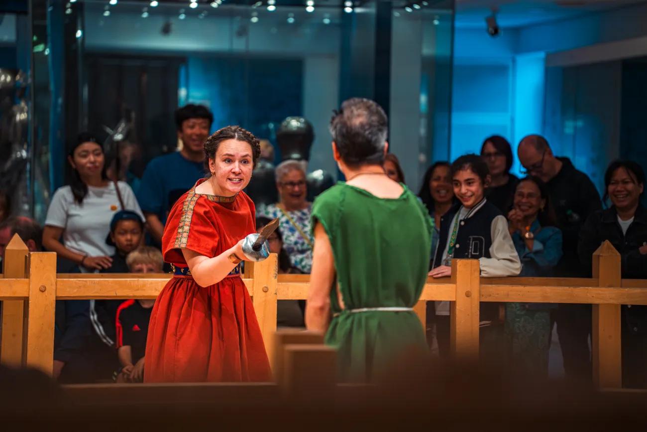 Two reenactors in red and green tunics perform a mock sword fight with wooden weapons inside a fenced area, while spectators observe from behind
