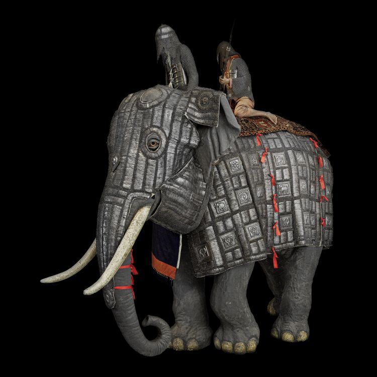 lifesize adult male elephant wearing armour with 2 riders on top